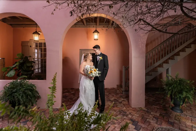 Alexis and Dino pose together under arched colonnade at The White Room, surrounded by potted greenery and hanging lanterns.