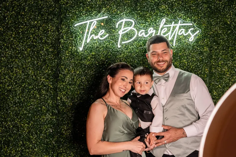 Alexis and Dino pose with a young child in front of a neon "The Barolittas" sign backed by green foliage.