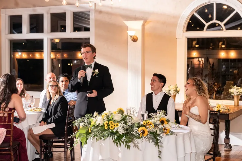 A man in a suit stands to toast while guests seated at a long table listen, with tall floral arrangements and lit windows