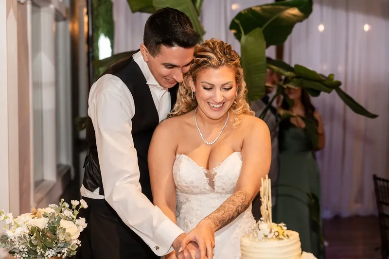 Dino and Alexis cut their wedding cake together as he leans toward her with greenery and white flowers flanking the dessert.