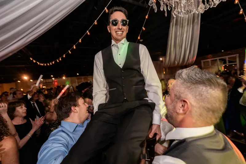 A man in a vest and sunglasses sits on guests' shoulders during the reception festivities.