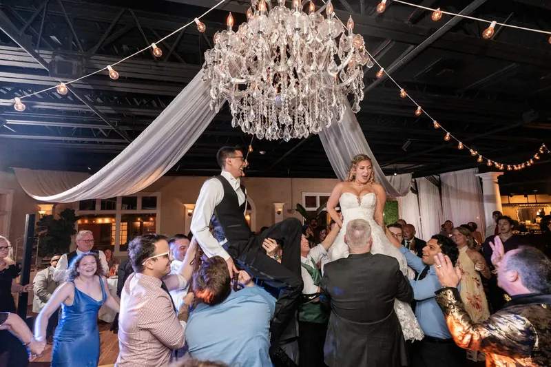 Alexis and Dino stand surrounded by cheering guests beneath the chandelier at their reception.