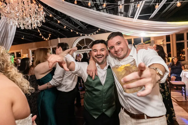 Dino grins while dancing with guests in a green vest at The White Room, surrounded by string lights and flowing white fabric.