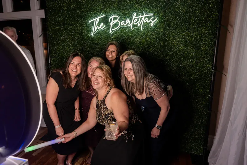 Four women pose together at a photo booth backdrop reading "The Barefoot's" against a lush green wall.