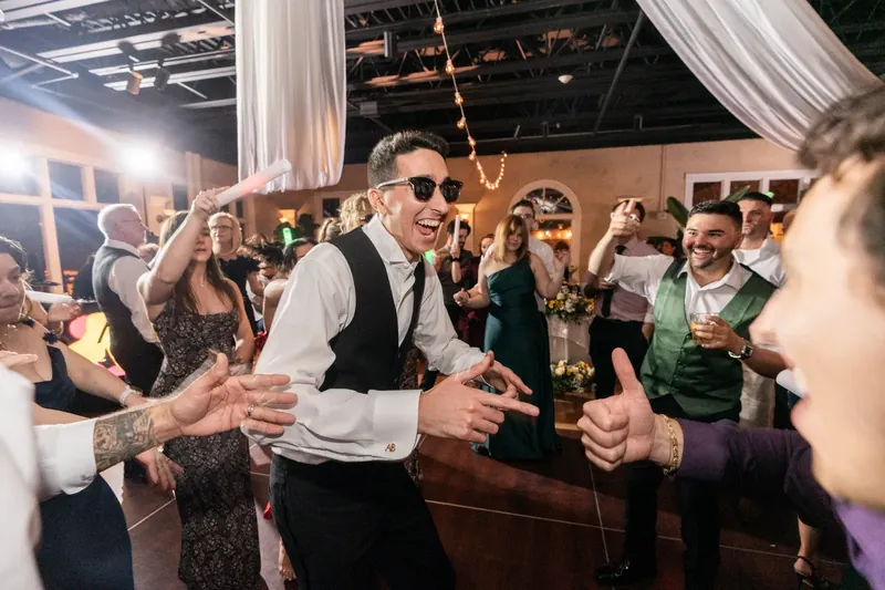 Dino dances energetically with guests during the reception, wearing sunglasses and a vest over his white shirt.