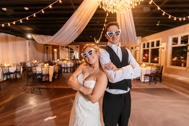 Alexis and Dino pose together wearing sunglasses in The White Room's illuminated ballroom, surrounded by chairs and warm