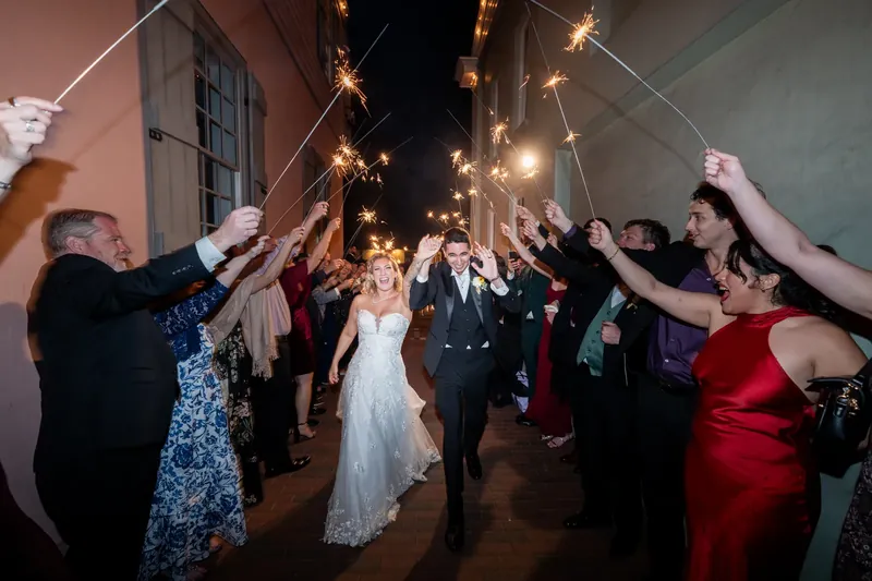 Guests raise sparklers high as Alexis and Dino walk between them down a glowing outdoor corridor at The White Room.