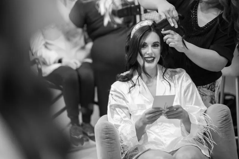 Black and white image of Alyson in a white robe seated while someone adjusts her hair from behind in the bridal suite.