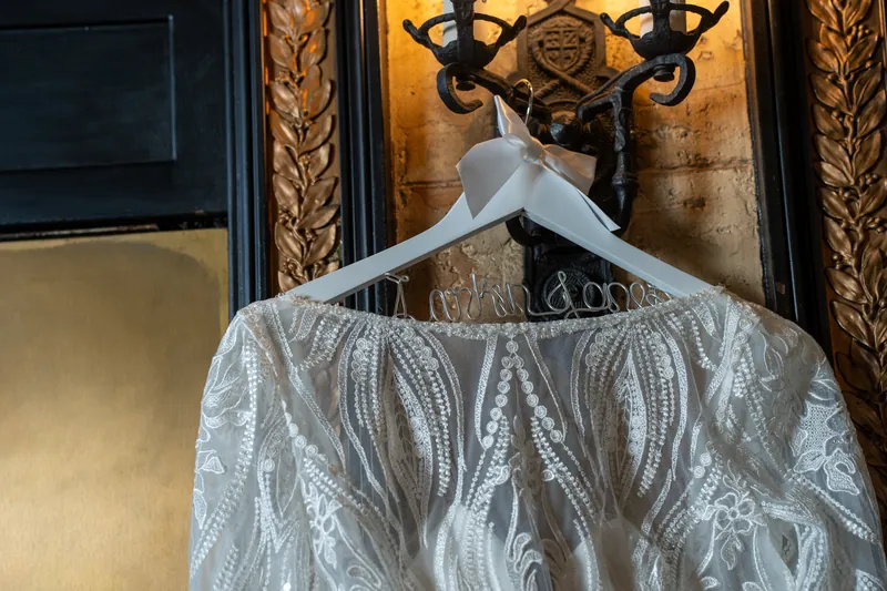A beaded white wedding dress hangs on a wooden hanger against ornate gold wall sconces at The Treasury on the Plaza.