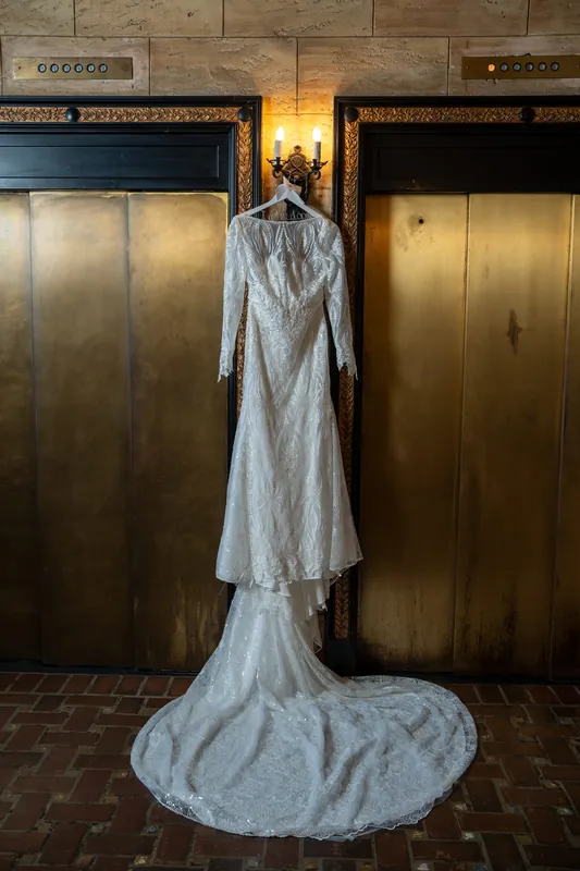 Alyson's lace gown with long train hangs between brass elevator doors at The Treasury on the Plaza.