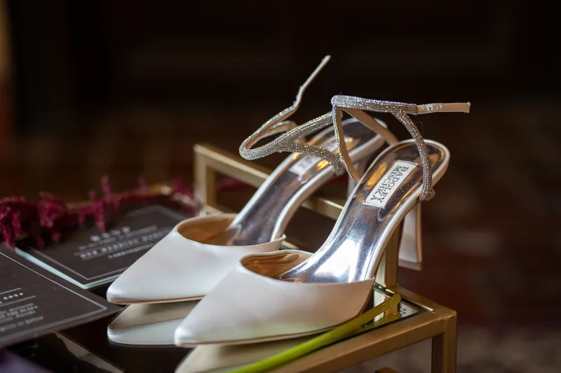 White satin heels with ankle straps sit on a gold-trimmed table near burgundy flowers and wedding details.