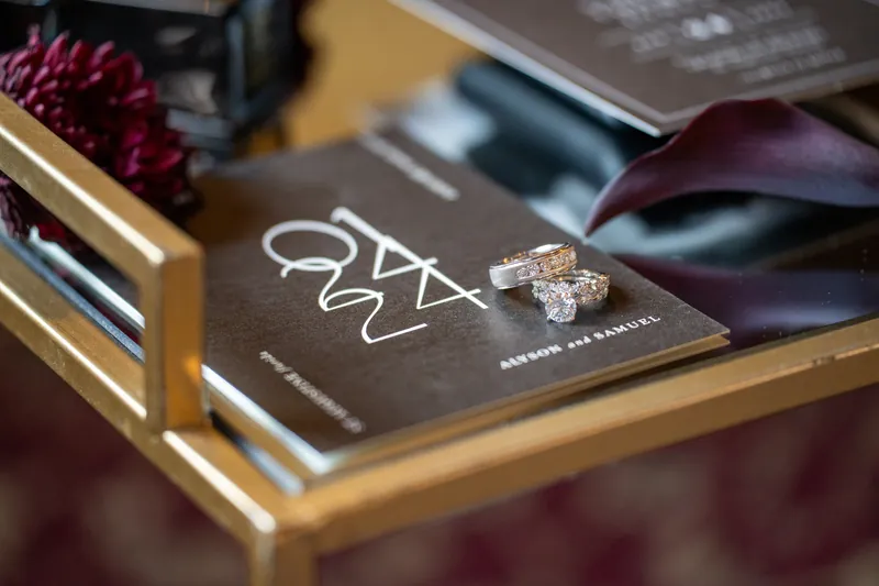 A wedding ring rests on a dark card with "Alyson & Sam" calligraphy, surrounded by burgundy flowers and wedding stationery.