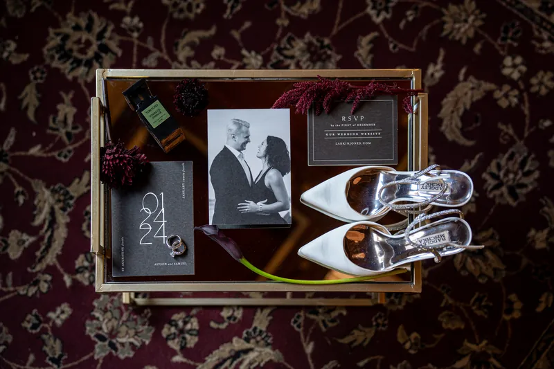 Wedding details arranged on burgundy damask include white heels, a black-and-white portrait, invitation, and keepsakes.