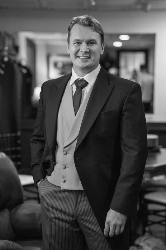 Sam smiles in a black suit with vest and burgundy tie against an indoor venue backdrop in black and white.