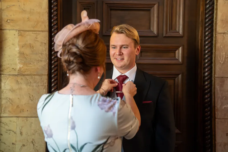 Someone fastens Sam's burgundy boutonniere while he stands at a wooden door in formal attire.