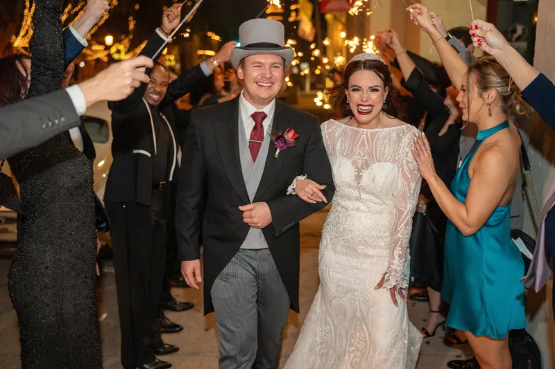 Alyson and Sam walk through a sparkler tunnel flanked by smiling guests in evening wear at their reception.