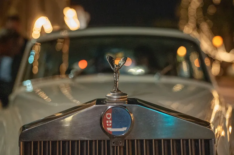 A golden hood ornament gleams on the vintage getaway car's hood, with twinkling lights blurred in the background.