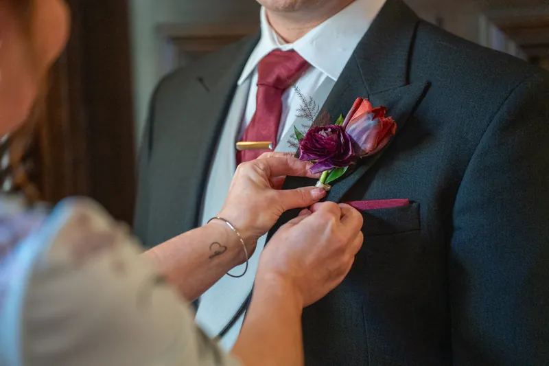 Hands pin a deep burgundy and purple floral boutonniere onto Sam's dark suit jacket.