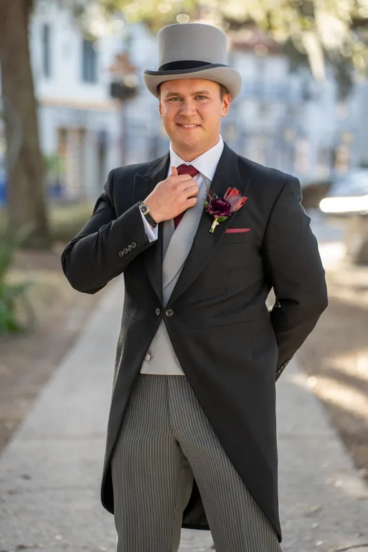 Sam adjusts his tie outdoors on a tree-lined St. Augustine street, wearing a gray morning coat and top hat.