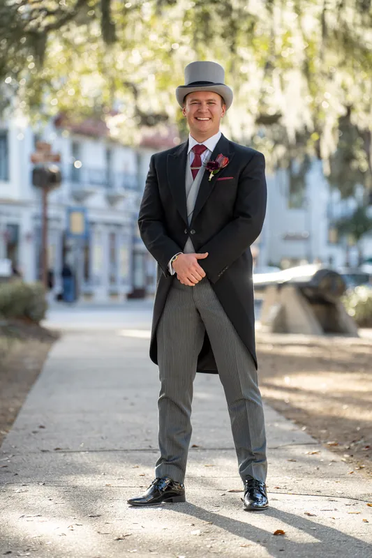 Sam stands in dappled sunlight on a brick pathway, one hand in his pocket, styled in formal morning attire.