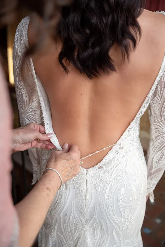 Hands fasten the back of a beaded white wedding dress, showing intricate embroidered detail and delicate buttons.
