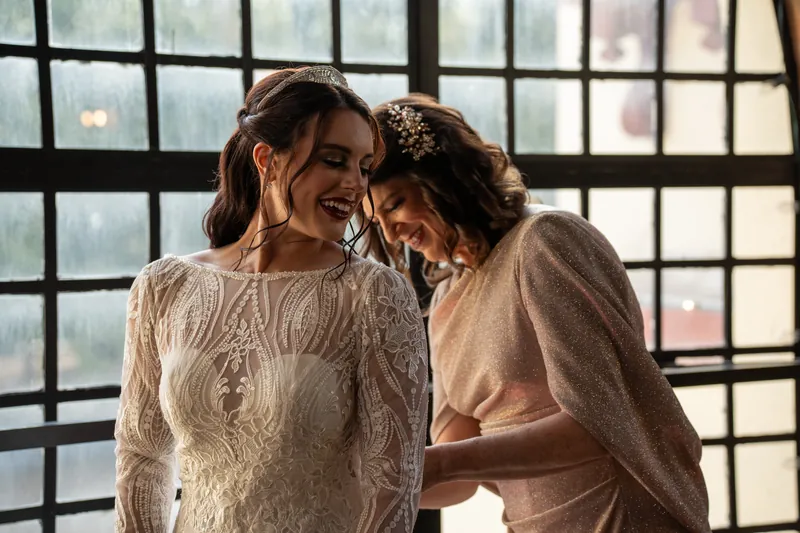 Alyson and a companion laugh together indoors near a window, both wearing cream-colored lace garments.