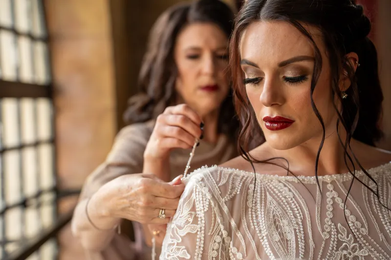 A friend helps secure Alyson's jewelry as she gazes downward, her dark wavy hair and burgundy lipstick visible in profile.