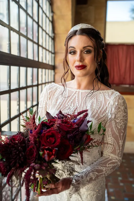 Alyson holds her dark crimson bouquet while wearing a beaded white gown, standing near a metal lattice window.