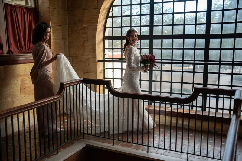 Alyson stands on a landing beside an arched window with gridded panes, holding her bouquet and looking toward a companion.