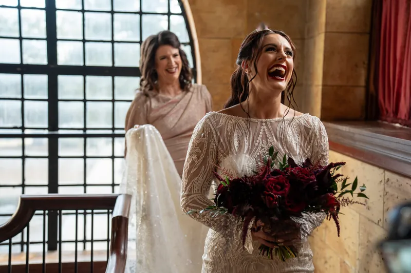 Alyson laughs with her dark bouquet while Sam stands behind her in an elegant stone room with tall windows.