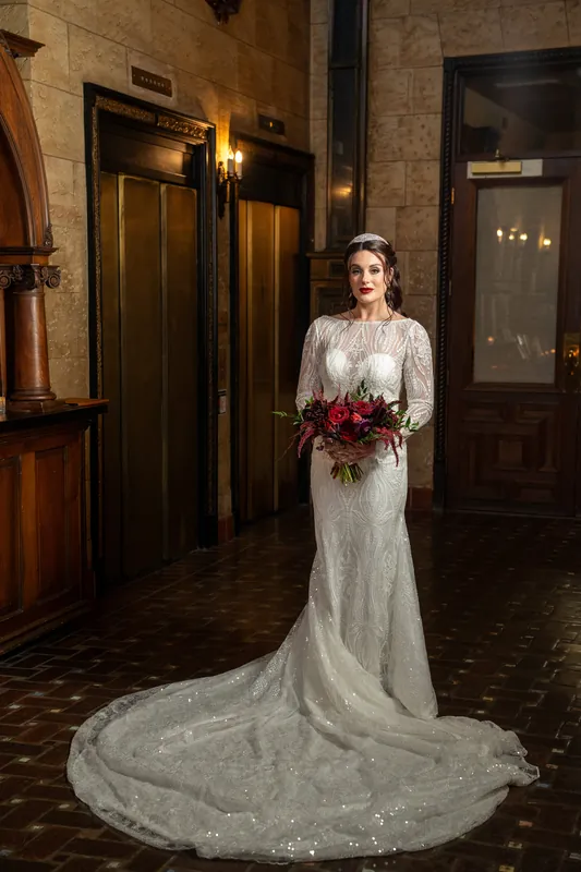 Alyson wears a flowing beaded gown with a dramatic train, holding her burgundy bouquet in a historic stone hallway.