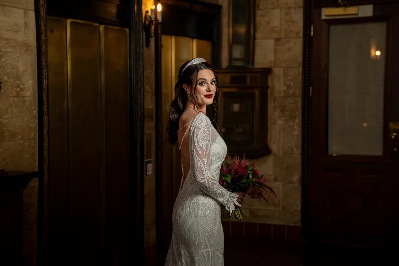 Alyson turns toward the camera in profile, her lace gown catching warm light from wall sconces, bouquet in hand.