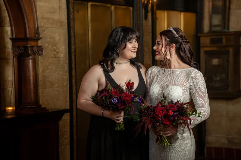 Alyson in black and Sam in cream face each other holding burgundy bouquets at The Treasury on the Plaza.