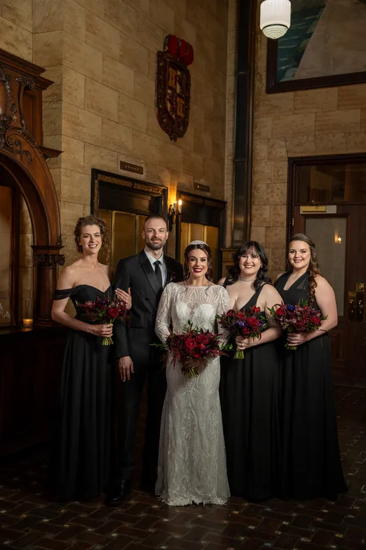 Sam and Alyson pose with five attendants in black dresses holding matching burgundy bouquets within the stone-walled venue.