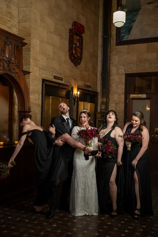 Alyson and Sam laugh with attendants holding deep red flowers beneath illuminated arches and sconces.