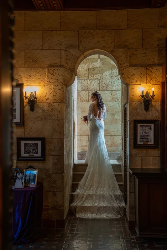 Alyson stands alone in a beaded white gown, veil trailing behind her, framed by stone archways and candlelit wall fixtures.
