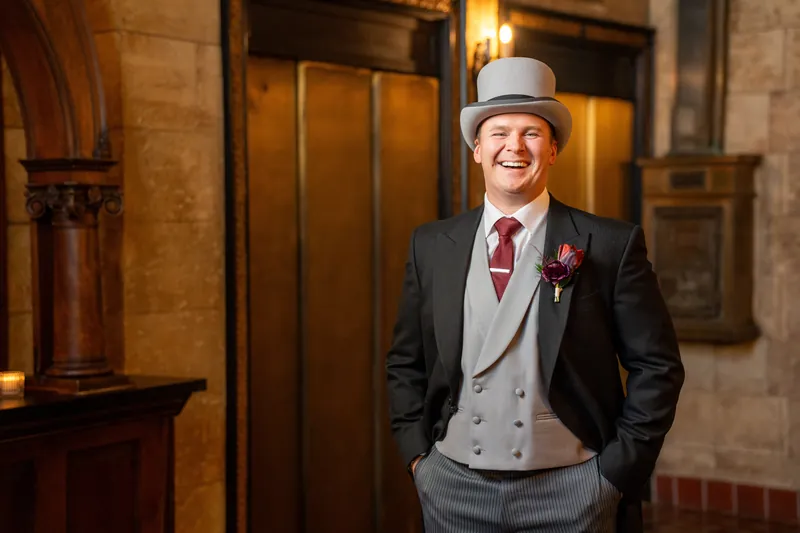 Sam wears a tan vest, charcoal suit, burgundy tie, and Panama hat in the stone-walled hall.