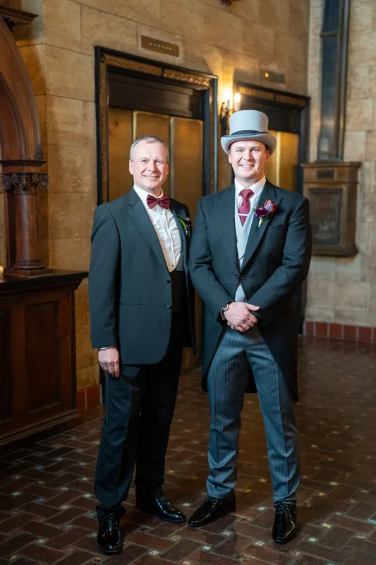Two guests in formal suits with boutonnières clasp hands in the historic stone corridor.