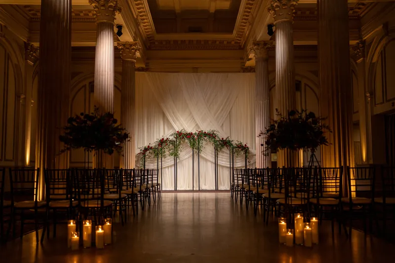 A floral arch with black wreaths and candlelit aisle fills the ornate ceremonial hall.