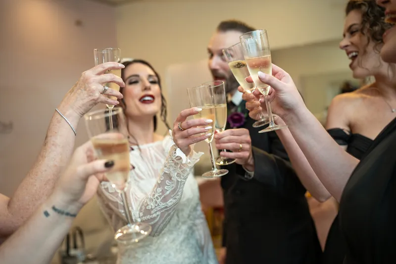 Alyson in a beaded white gown toasts with champagne alongside two smiling guests.