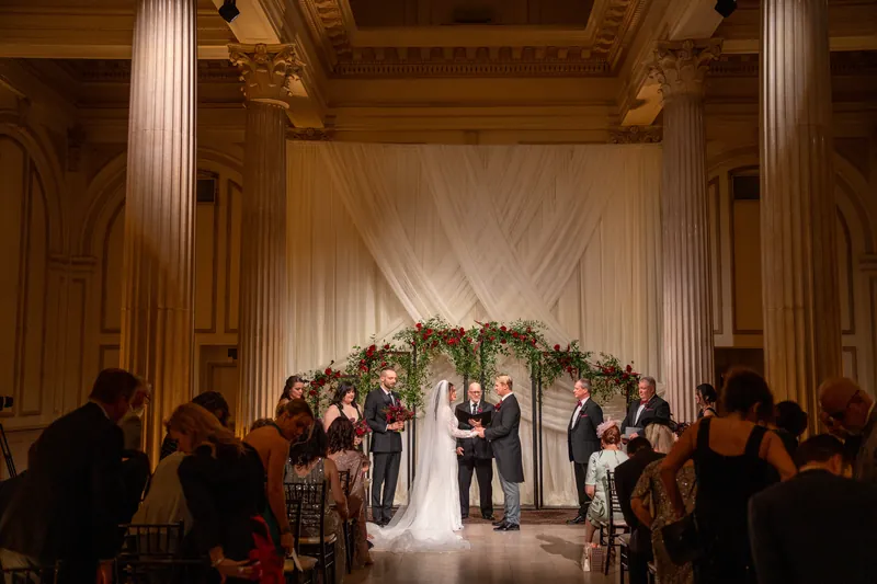 The couple stands beneath a draped floral arch as guests watch from wooden chairs at The Treasury on the Plaza.