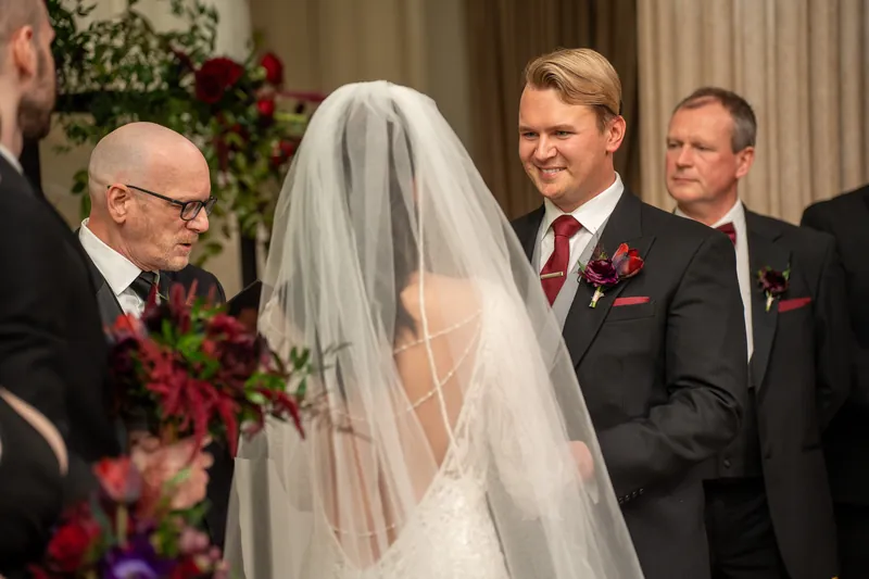 Sam smiles at Alyson as she faces the officiant under the decorated arch at The Treasury on the Plaza.