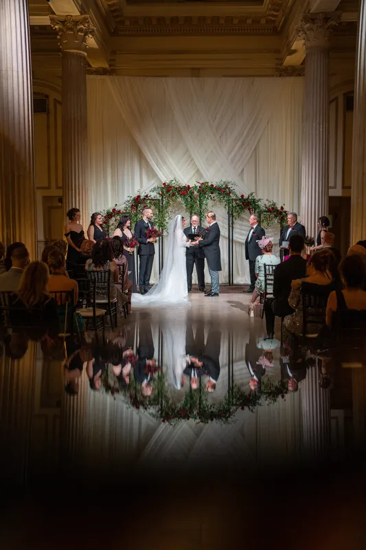 Alyson and Sam exchange vows beneath a lush floral arch at The Treasury on the Plaza, reflected in the polished floor below.