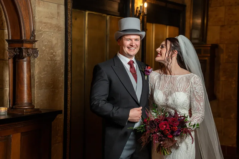 Sam wears a top hat and burgundy tie while Alyson holds her bouquet; both smile at each other in the stone-walled interior.