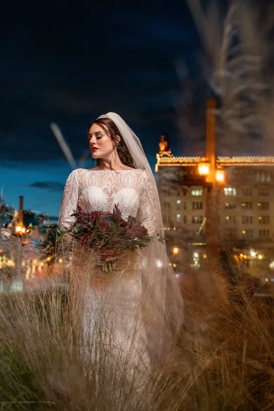 Alyson in a beaded lace gown and veil holds her bouquet while gazing to the side with lit buildings behind her at dusk.