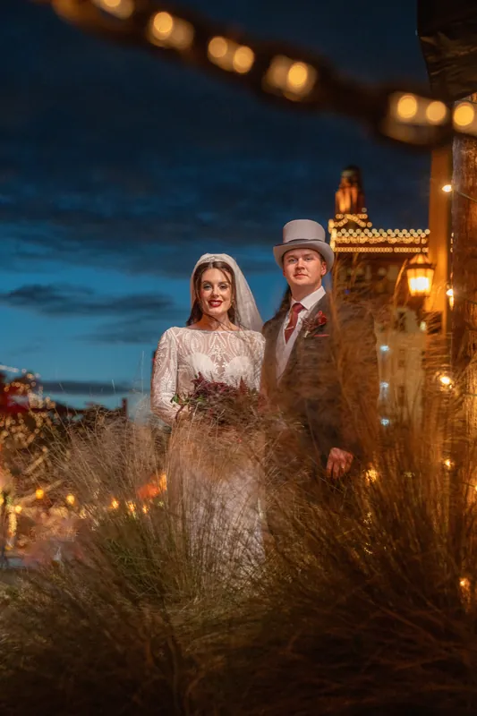Sam and Alyson stand outdoors at dusk with glowing string lights and warm brick buildings behind them.