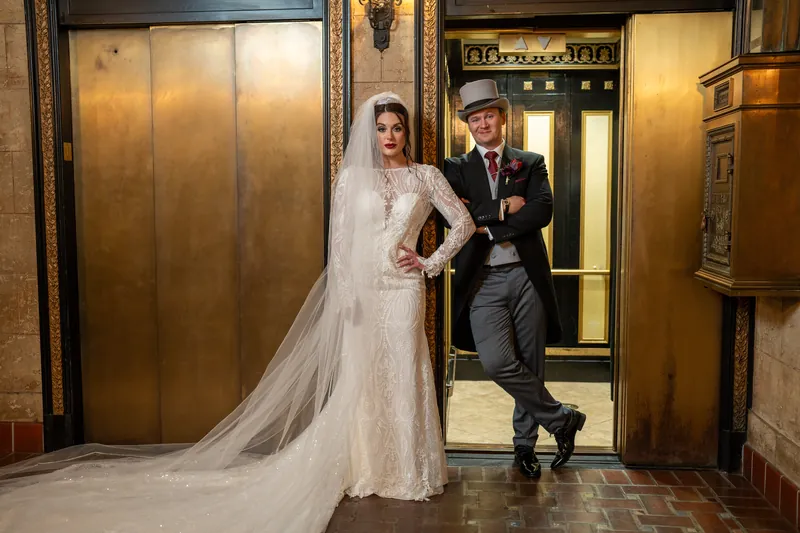 Standing in the ornate elevator doorway, Alyson displays her beaded gown while Sam adjusts his jacket.