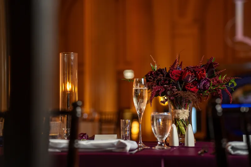 A reception table features a dark floral arrangement with burgundy blooms, candlelight, and place settings.