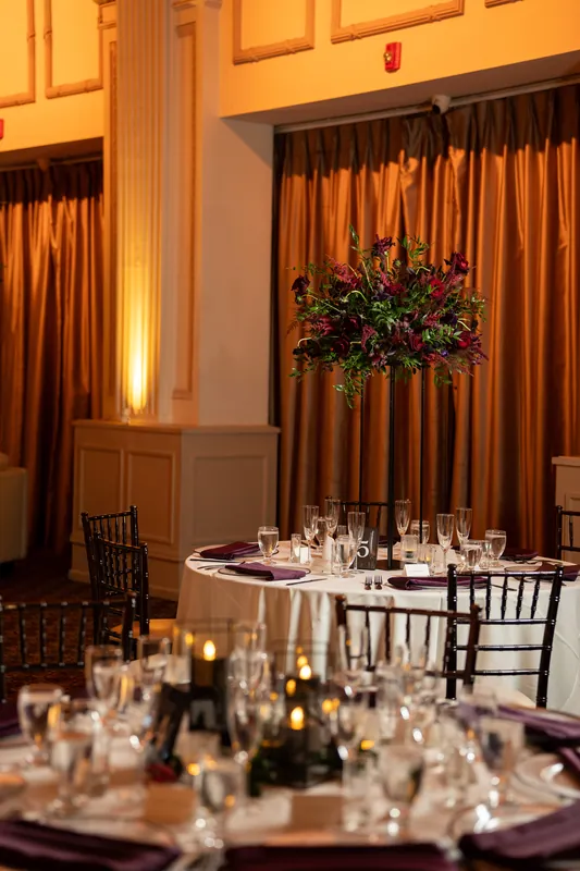 Round tables set with linens and glassware sit beneath gold curtains and a suspended floral arrangement.