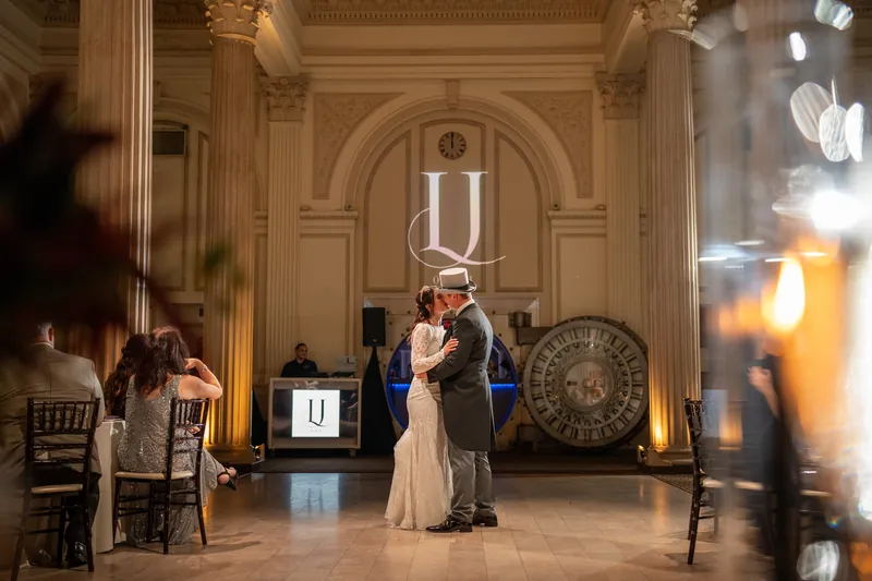 Alyson and Sam hold each other close on the dance floor beneath architectural columns and warm lighting.